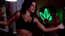 a woman in a black crop top is dancing in a room with a neon sign in the background .