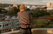 a woman in a striped sweater stands on a balcony overlooking a city with the word offspring on the bottom right