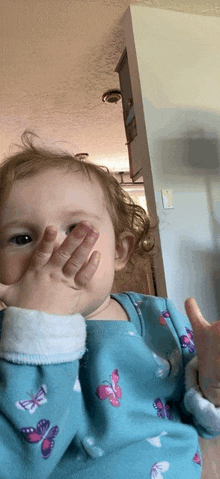 a baby wearing a blue butterfly pajama covering her face with her hands