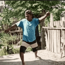 a man in a blue shirt and white shorts is standing in front of a wooden fence with his arms outstretched .