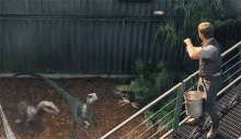 a man is standing on a balcony with a bucket in front of a fence with two dinosaurs in the background .