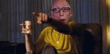 a man with glasses is sitting in a chair holding a remote control and pointing at the camera .