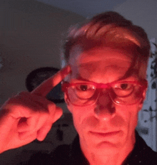 a man wearing red glasses holds his finger to his head