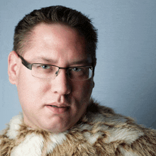 a man wearing glasses and a fur coat has a serious look on his face