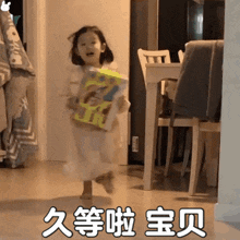 a little girl in a white dress is running in a room holding a box with chinese writing on it