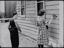 a man and a woman are standing in front of a house . the woman is painting a heart on the side of the house .