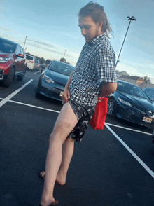 a man in a plaid shirt holds a red bag in a parking lot with cars