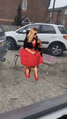 a woman in a red dress sits on a pink bench with a car parked behind her