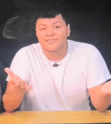 a man in a white t-shirt is sitting at a table and making a gesture with his hand .