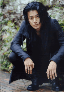 a man with long black hair is squatting down on a brick sidewalk