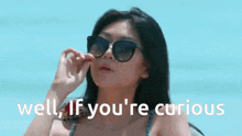 a woman wearing sunglasses is standing on a beach with the words well if you 're curious behind her .