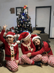 three children wearing santa hats are sitting on the floor in front of a christmas tree