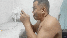 a shirtless man is sitting in front of a fan and looking at his phone