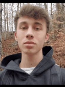 a young man wearing a black hoodie stands in front of trees