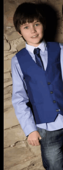 a boy wearing a blue vest and tie is standing in front of a stone wall