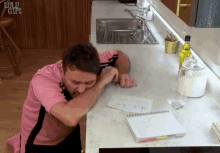 a man in a pink shirt sits on a kitchen counter with a bottle of olive oil in the background