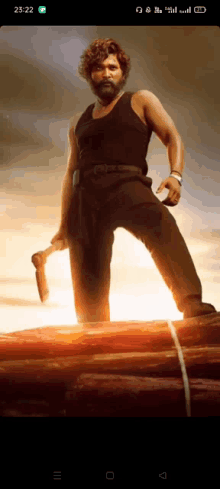 a man in a black tank top is holding an axe and standing on a pile of logs