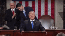 a man in a suit and tie is sitting at a podium giving a speech while two other men stand behind him .