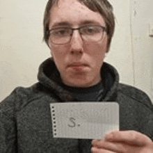 a young man wearing glasses is holding a piece of paper with the word s. on it .