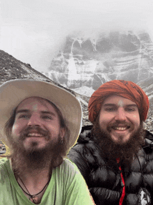 a man with a beard wearing a hat and a scarf with a mountain in the background smiles for the camera