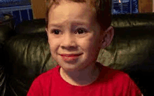 a young boy in a red shirt is sitting on a couch and making a funny face .