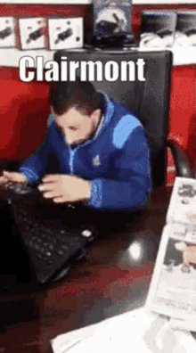 a man in a blue jacket is sitting at a desk in front of a laptop computer with the name clairmont written above him .