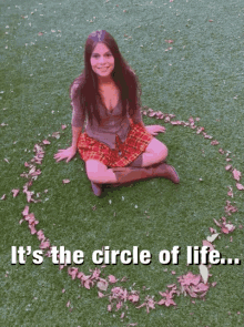 a woman in a plaid skirt sits on the grass with her arms outstretched and a circle of leaves around her knee