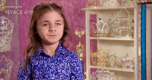 a young girl in a blue shirt is standing in front of a shelf filled with trophies and tiaras .