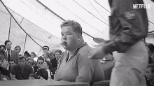 a black and white photo of a man sitting at a table in front of a crowd of people .