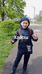 a young boy wearing a blue jacket and a backpack is standing on a sidewalk and giving a peace sign .