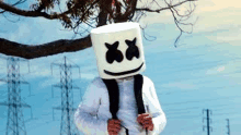 a man wearing a marshmallow mask and a backpack is standing in front of a tree .