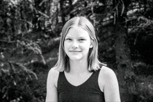 a girl in a black tank top stands in front of trees