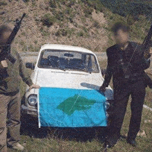 two men holding guns are standing in front of a car and a flag .