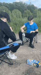 two boys are sitting in folding chairs on a dirt road and one of them is pouring water into a bottle