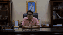 a man in a pink shirt sits at a desk in front of a framed picture of a man in a white robe