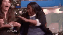 a woman is sitting in front of a christmas tree and laughing while another woman hugs her .