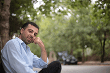 a man in a blue shirt leans against a tree with his hand on his chin