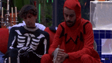 two men dressed in skeleton and devil costumes sit on a couch