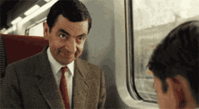 a man in a suit and tie is sitting on a train and looking at another man .