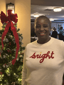 a woman wearing a white sweater that says bright is smiling in front of a christmas tree