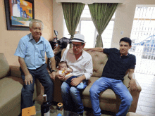 three men are sitting on a couch holding a baby and one of them has a hat on