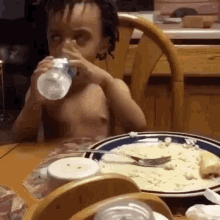 a young boy is drinking water from a bottle while sitting at a table with a plate of food ..