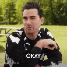 a man in a black and white jacket is sitting at a picnic table with his hand on his knee and says okay .