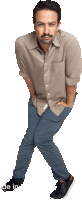 a man in a tan shirt and blue pants stands with his hands in his pockets on a white background