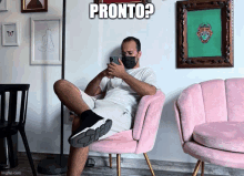 a man wearing a mask is sitting in a pink chair looking at his phone with the caption pronto