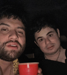 a man with a beard holds a red cup next to another man