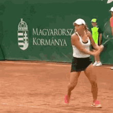 a woman is swinging a tennis racquet on a tennis court in front of a banner that says magyarország kormánya .