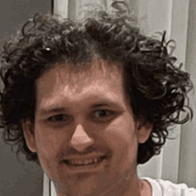 a man with curly hair and a white shirt is smiling for the camera .