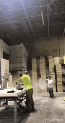 a man in a green shirt throws a pizza in a warehouse with a stack of wooden pallets in the background