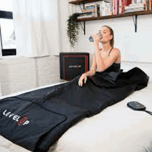 a woman is drinking water from a bottle while laying in a black sleeping bag on a bed .